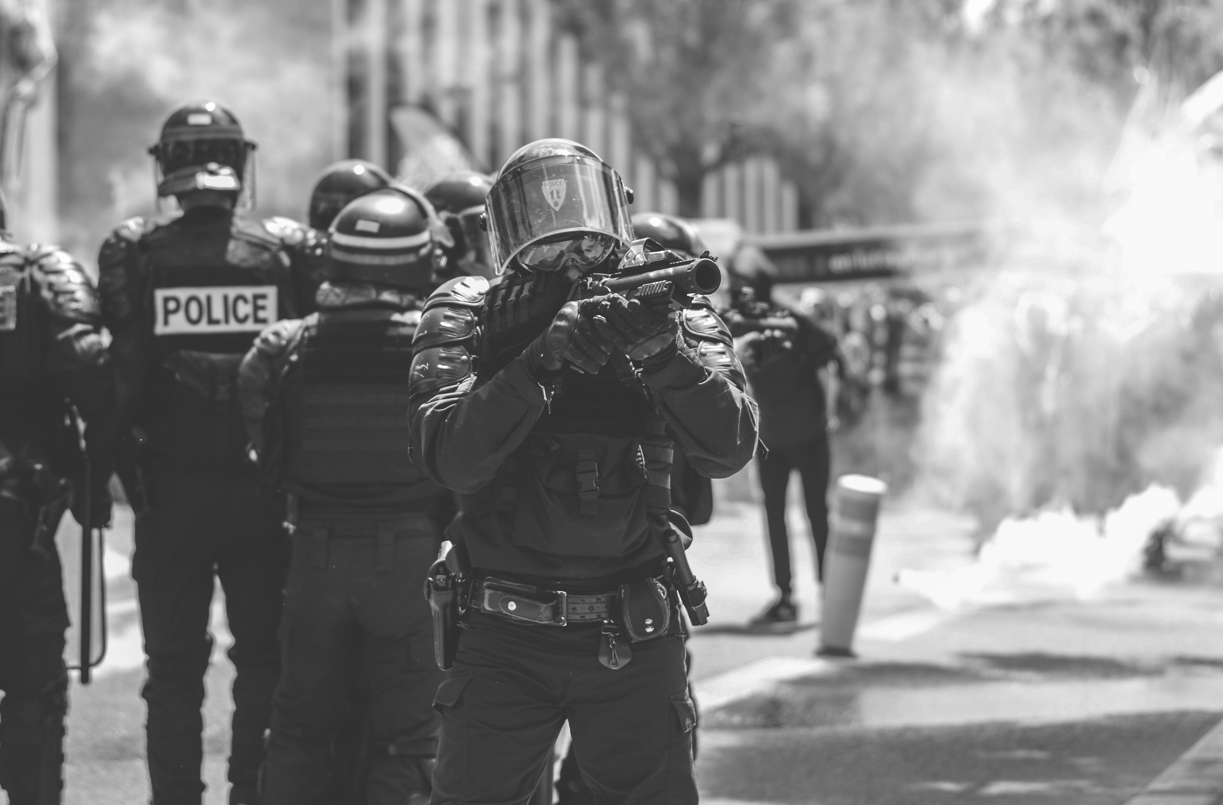 Riot police amid smoke, with a foreground officer aiming a launcher